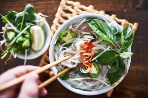 Top-down view of a bowl of Vietnamese pho with fresh herbs, lime, and a spicy sriracha drizzle, held with chopsticks.